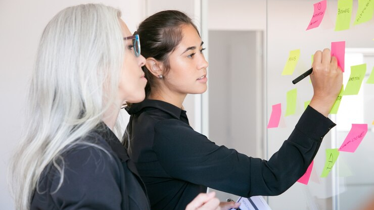 Two women brainstorming with sticky notes on a glass board, representing innovative marketing trends for 2025.