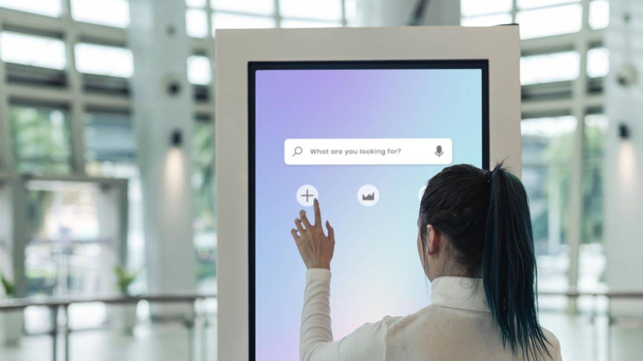A woman interacting with a digital touch screen featuring an AI-powered search engine with voice and text search capabilities.