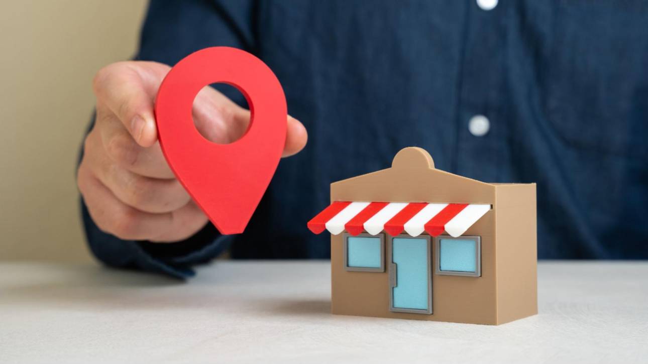 Hand holding a red location pin next to a small store building for Google Business Profile sync.