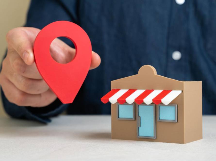 Hand holding a red location pin next to a small store building for Google Business Profile sync.